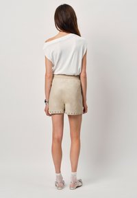Beige shorts met studs aan de zoom en een elastische tailleband, gecombineerd met een witte, off-shoulder top. Het model staat met de rug naar de camera, waardoor de outfit goed zichtbaar is.