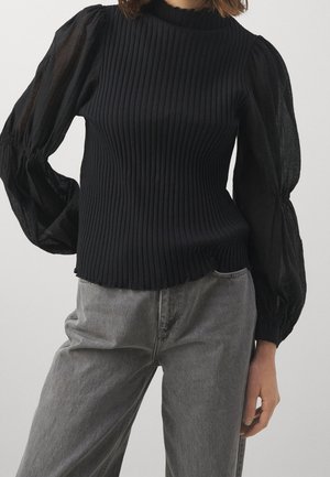 Kvinde iført en sort ribbet sweater med gennemsigtige ballonærmer og grå jeans med højtaljede, stående mod en ensfarvet baggrund.