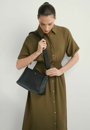 Kobieta w oliwkowej sukience na guziki, trzymająca czarną skórzaną torebkę na ramię z grubym paskiem na tle jasnego, jednolitego tła.