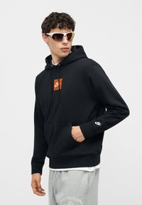 Νέος άνδρας που φοράει μαύρη μπλούζα Nike με πορτοκαλί λογότυπο, γκρι φόρμες, λευκά γυαλιά ηλίου, ποζάροντας με το ένα χέρι στην τσέπη μπροστά σε λευκό φόντο.