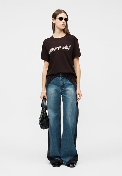 Γυναίκα που φοράει μαύρο γραφικό μπλουζάκι, τζιν παντελόνι με φαρδιά μπατζάκια σε δύο τόνους μπλε και μαύρο, μαύρα παπούτσια, στρογγυλά γυαλιά ηλίου, κρατώντας μαύρη τσάντα στο πλάι.