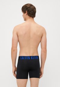 Boxeri negri dintr-un material neted și elastic, cu un brâu logo "CALVIN KLEIN" de culoare albastră. Au un design ajustat și spate fără cusături.