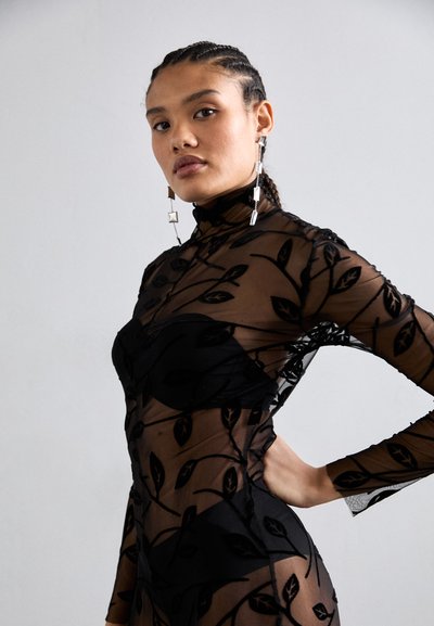 Black sheer mesh bodysuit with leaf patterns, high neckline, long sleeves, and fitted design. Large geometric earrings complement the look.