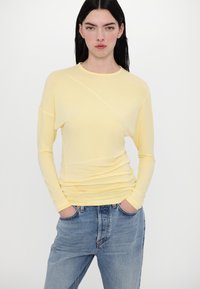 Gelbes Langarmshirt aus leicht strukturiertem Stoff, mit Rundhalsausschnitt und Raffung, kombiniert mit blauen Denim-Jeans.