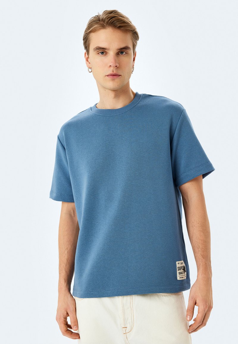 Blå bomulls t-shirt med korta ärmar, rund halsringning, och en etikett på nederkant till höger. Tyget ser mjukt ut med en slät yta.