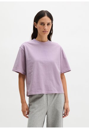 Kobieta z długimi ciemnymi włosami, ubrana w luźną lawendową koszulkę z krótkim rękawem i jasnoszare spodnie, stojąca na tle gładkiego białego tła.