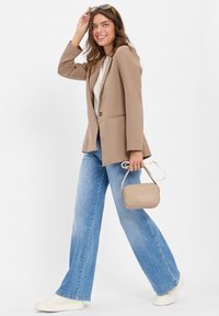 Lichtbeige blazer, witte top en lichtblauwe, hoge taille, wijde jeans. Houdt een kleine beige handtas vast. Witte sneakers maken de look compleet.