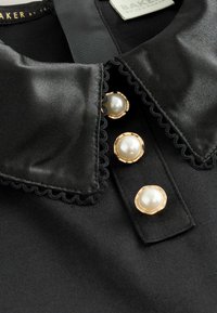 Melna auduma bluze ar satīna apvalku, dekorēta ar trim zeltainiem pērļu pogām un viļņotām malām. Ziņojums redzams apkaklītes daļā.