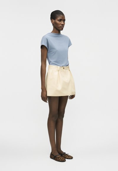 Γυναίκα όρθια, φοράει ανοιχτό μπλε μπλουζάκι με κοντά μανίκια, μίνι φούστα κρεμ με ζώνη και φλατ παπούτσια με λεοπάρ σχέδιο, πάνω σε απλό φόντο.