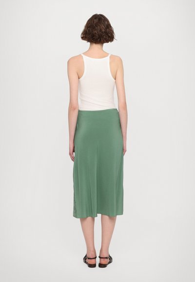 Groene midi-rok met een gladde textuur, voorzien van een kanten detail aan de zijkant. Gecombineerd met een aansluitend wit mouwloos shirt en sandalen.