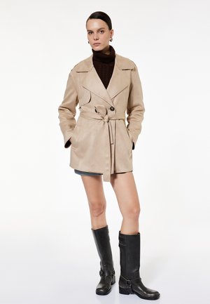 Mujer con las manos en los bolsillos que lleva un abrigo trench beige con cinturón, un suéter oscuro de cuello alto, falda corta y botas negras hasta la rodilla.