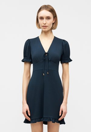 Kobieta ubrana w krótką granatową sukienkę z bufkami, dekoltem w serek z wiązaniem oraz falbaną na dole, stojąca na jednolitym tle.