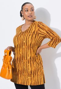 Gelb-orange Tunika mit braunen gemusterten Streifen, V-Ausschnitt, Dreiviertelärmel und seitlichen Taschen. Kombiniert mit einer orangefarbenen Zugtasche.