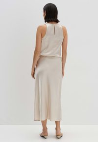 Robe sans manches beige clair avec un détail en forme de trou de serrure dans le dos, texture lisse et jupe mi-longue. Associée à des chaussures slip-on beiges.