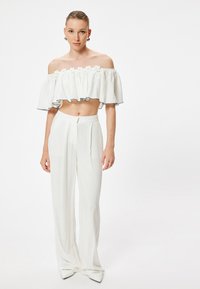 Crop top bianco con spalle scoperte, orlo a balze e dettagli floreali, abbinato a pantaloni bianchi a vita alta con gamba dritta.