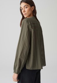 Blusa trasparente verde oliva a maniche lunghe con strisce verticali e dettagli di cucitura sottili. Vestibilità ampia con polsini con bottoni e orlo arrotondato.