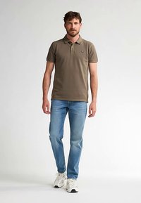 Khaki-Poloshirt mit kurzen Ärmeln und kleinem Logo. Hellblaue Jeans, schmal geschnitten, mit verwaschenem Design. Weiße Sportschuhe mit schwarzen Akzenten.