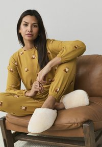 Geltona pižama su lapės atspaudu, su elastinėmis rankovėmis. Baltos pūkuotos šlepetės ant rudos odinės kėdės, pabrėžiančios minkštus tekstūras ir kontrastą.