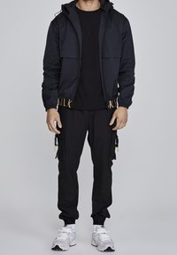 Black bomber jacket with a hood, zip front, and elastic cuffs, paired with a black t-shirt and cargo pants with pockets. White sneakers.