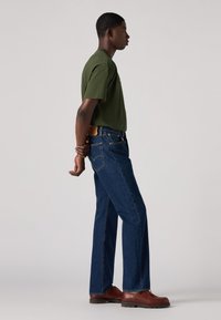 Joven de pie de lado con las manos entrelazadas detrás de la espalda, vistiendo camiseta verde, jeans azules y zapatos de cuero marrón, contra un fondo liso.