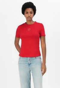 T-shirt rouge ajustée avec un col rond et des manches courtes, arborant un logo circulaire en blanc. Associée à un jean en denim délavé.