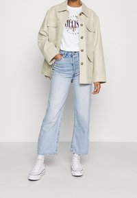 Person wearing beige buttoned jacket over white graphic t-shirt, light blue high-waisted straight jeans, and white high-top sneakers.