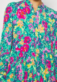 Abito floreale con uno sfondo turchese vibrante e fiori rosa, gialli e verdi. Include un colletto con bottoni e un design della gonna plissettata.