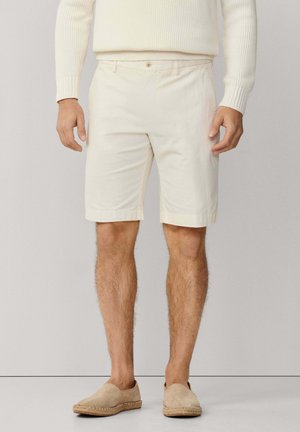 Hombre que lleva pantalones cortos color crema hasta la rodilla, alpargatas beige claro y un suéter de canalé color crema, de pie contra un fondo liso.