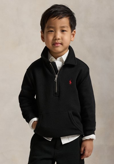 Jeune garçon portant un pull noir à demi-fermeture éclair avec un logo rouge et une chemise blanche en dessous, debout devant un fond neutre uni.