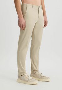 Pantalones beige claro de corte slim, con cremallera y bolsillos delanteros. La tela parece suave y con un ligero estiramiento. Combinados con zapatillas ligeras.