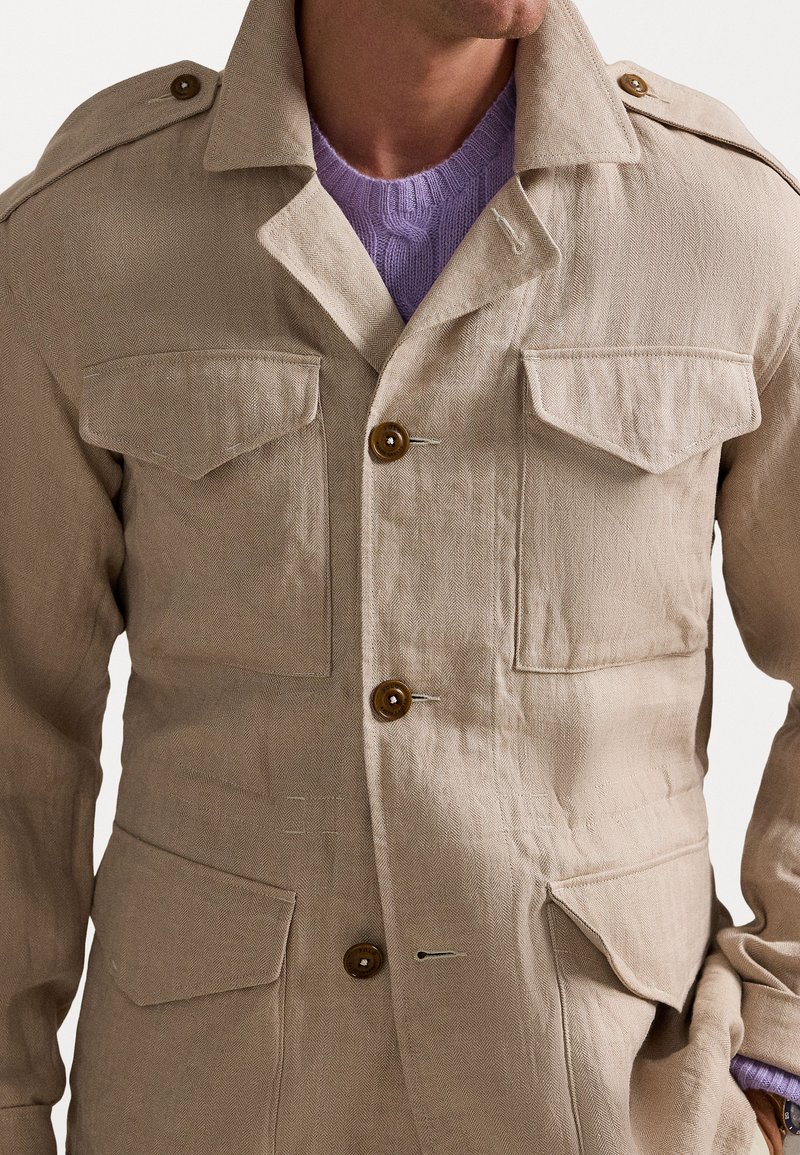 Veste en lin beige à texture chevrons, avec deux poches poitrine et des boutons marron. Portée par-dessus un pull en tricot violet clair.