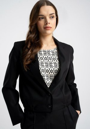 Kobieta z długimi brązowymi włosami, ubrana w czarną marynarkę i czarno-białą wzorzystą bluzkę, stojąca z rękami w kieszeniach na jednolitym tle.