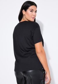Camiseta negra de manga corta con cuello redondo, vista desde la parte posterior. La tela parece suave y ligera, combinada con pantalones negros brillantes.
