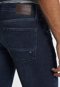 Jeans in denim blu scuro con patch del marchio in pelle, cuciture sulla tasca posteriore e piccolo dettaglio con logo tricolore nell'angolo della tasca.