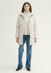 Manteau matelassé crème avec capuche, doté d'une fermeture éclair, d'un design matelassé et de poches latérales, associé à un pull en tricot épais et à un jeans bleu.