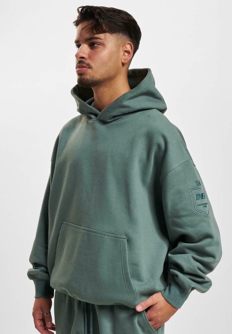 Un hoodie ample vert en tissu doux, avec une poche kangourou, des poignets côtelés et un détail de logo sur la manche.
