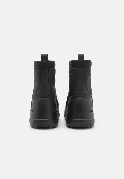 Bottes noires à la cheville avec un dessus synthétique lisse, un poignet élastique et une semelle moulée. Détail du logo distinct sur le côté.