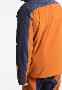Veste bicolore avec une partie supérieure bleu marine et une partie inférieure orange. Fabriquée en tissu lisse et léger, équipée d'une capuche.