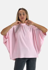 Rosa, locker sitzende Bluse mit hohem Ausschnitt und kurzen, weiten Ärmeln. Strukturierter Stoff mit subtilen Drapierungselementen. Kombiniert mit dunklen Jeans.