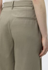 Κοντινό πλάνο μπεζ ραμμένων παντελονιών που δείχνει την πίσω τσέπη, τις θηλιές για ζώνη και το δεξί χέρι ενός ατόμου που ακουμπάει στο πλάι τους.