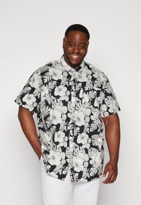 Camicia a maniche corte nera con un motivo floreale bianco, dotata di colletto, chiusura con bottoni e vestibilità relax, abbinata a pantaloni bianchi.