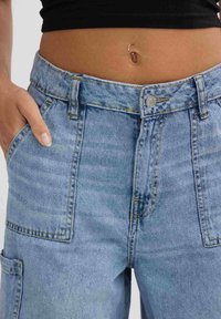 Pantalones cargo de denim azul claro con cintura alta, múltiples bolsillos y detalles de costura visibles. La tela tiene una textura ligeramente desgastada.