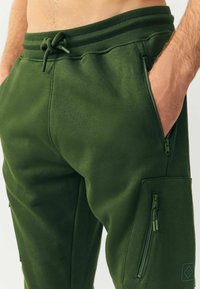 Grüne Jogginghosen aus weichem, dehnbarem Stoff, mit einem Kordelzugbund, seitlicher Reißverschlusstasche und strukturierten Akzenten.