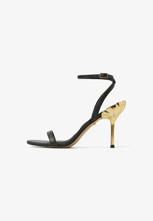 Sandalias de tacón negro con una delgada correa en el tobillo y plantilla texturizada. El tacón es de un dorado brillante con un diseño curvado y punta afilada.