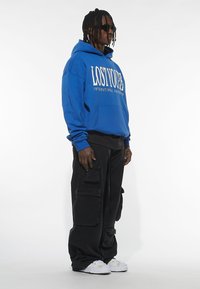 Plava dukserica s logom "LOST YOU", u kombinaciji s širokim crnim cargo hlačama s više džepova. Bijele tenisice upotpunjuju outfit.