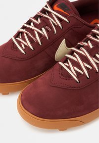 Μπορντό αθλητικά παπούτσια με σουέντ φινίρισμα, μπεζ κορδόνια και ταμπά λαστιχένια σόλα. Διαθέτουν έντονο μπεζ λογότυπο Nike στο πλάι.