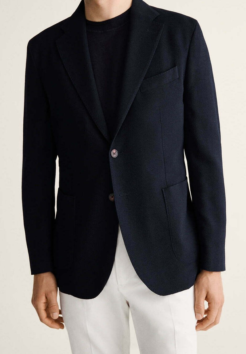 Hombre que lleva una chaqueta negra sobre una camisa negra combinada con pantalones blancos sobre un fondo neutro.