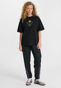 Czarna bawełniana koszulka z krótkim rękawem i zielono-białymi haftami. W zestawie z czarnymi spodniami, które mają szare boczne panele i elastyczne mankiety.