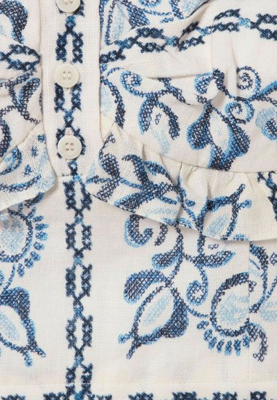 Tessuto bianco con ricamo floreale blu, quattro bottoni bianchi in linea verticale e un piccolo dettaglio arricciato vicino alla parte superiore destra.