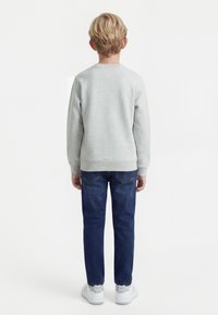 Blonds bērns stāv ar muguru pret skatītāju, valkājot gaiši pelēku sporta krekliņu, tumši zilas džinsas un baltus sporta apavus uz vienkārša fona.